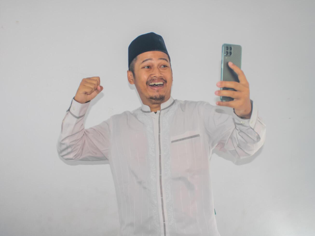 Moslem Asian man clenched fist showing excitement while looking to his mobile phone photo