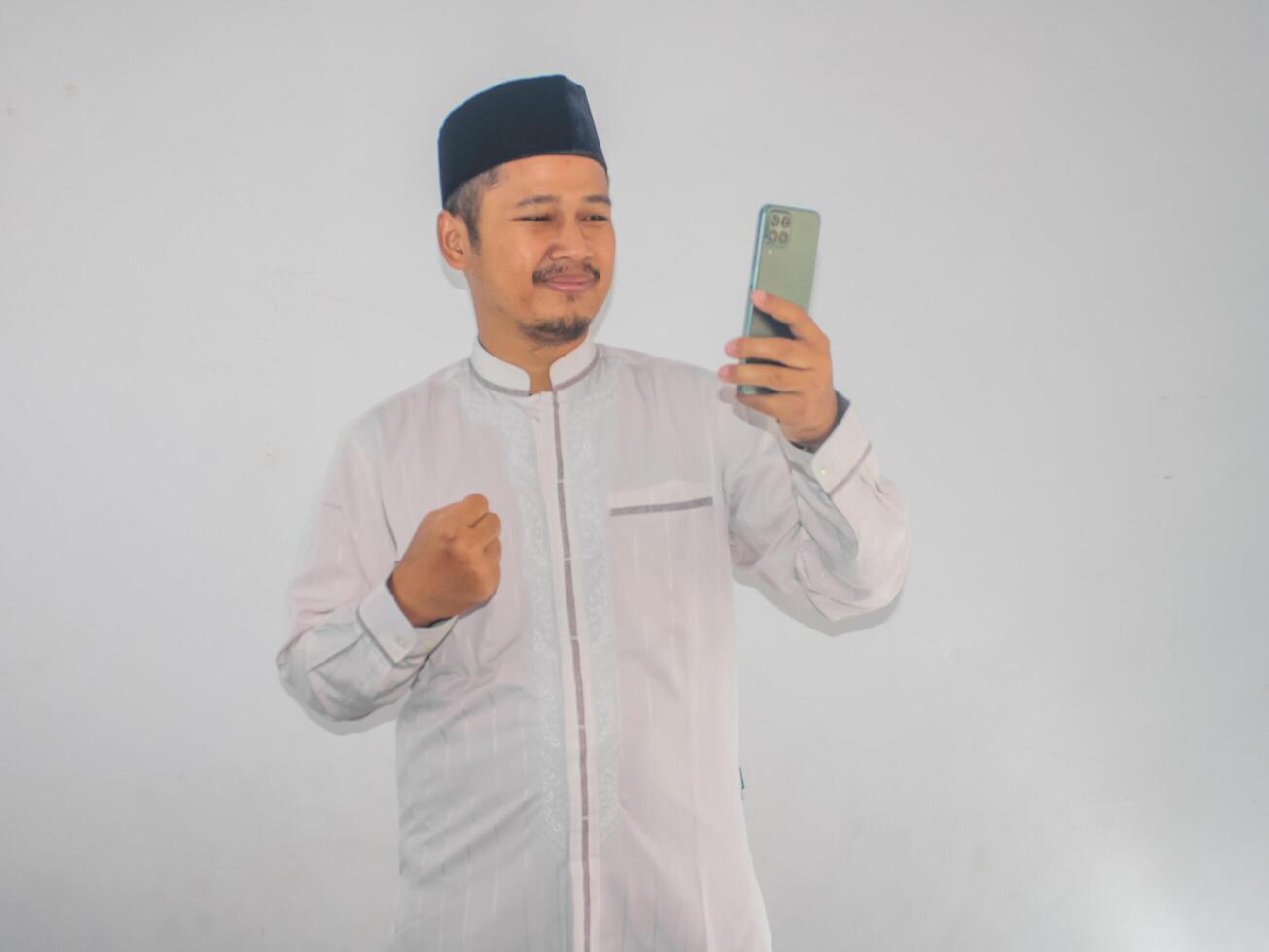 Moslem Asian man clenched fist showing excitement while looking to his mobile phone photo