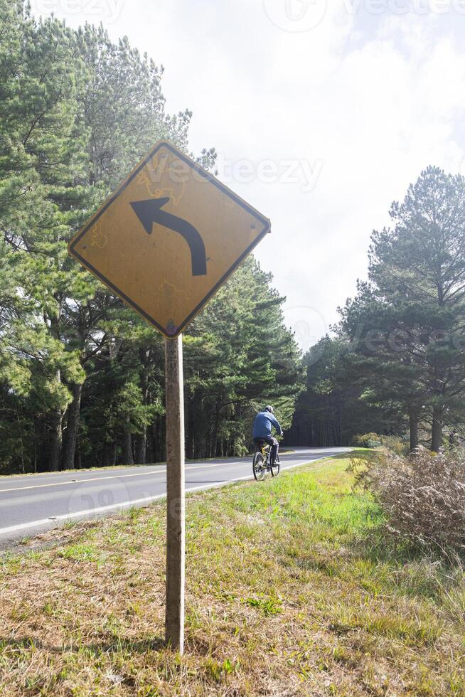 a road in the middle of the trees with a signpost and a man riding a bicycle in the background photo