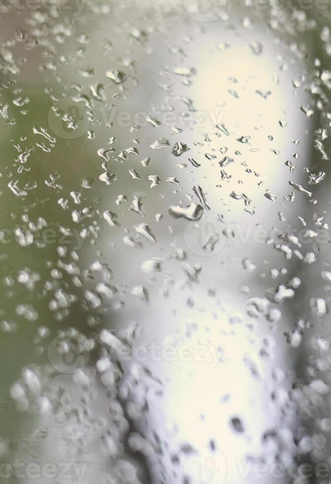 A photo of rain drops on the window glass with a blurred view of the blossoming green trees. Abstract image showing cloudy and rainy weather conditions