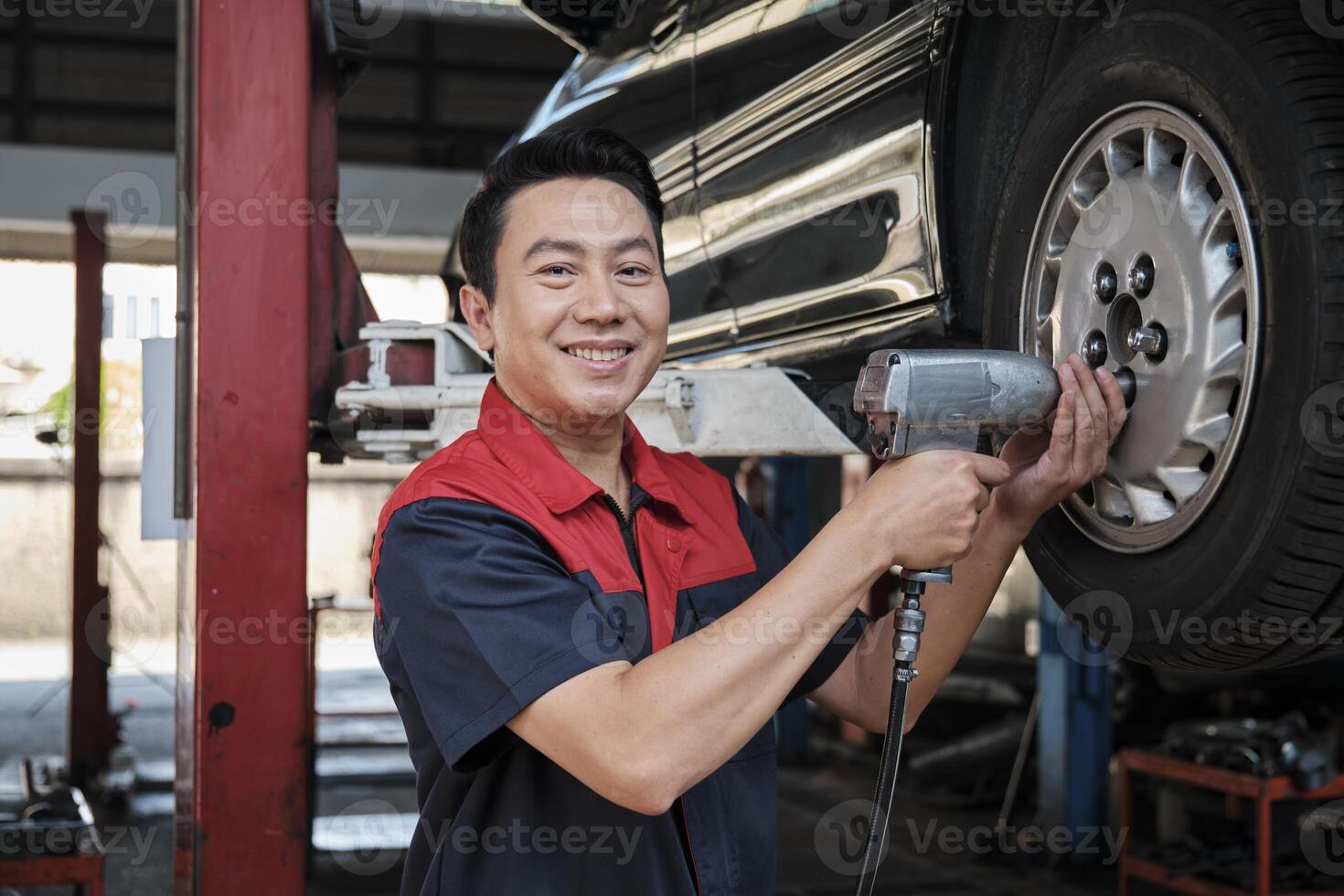 One expert Asian male automotive mechanic technician is screwing car wheel nuts on lifting with drill wrench for repair at garage. Vehicle maintenance service works industry occupation business jobs. photo