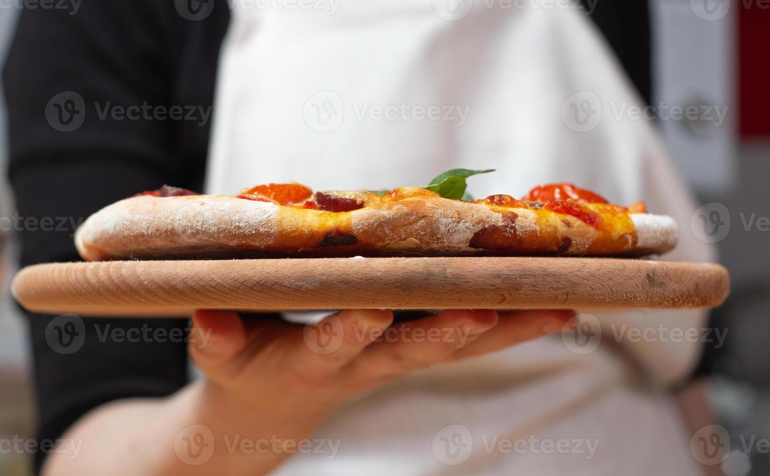 Chef holds wooden tray or board with homemade cheese pizza with tomato cherry, sauce, sausage and basil. Tasty dinner, close up view photo