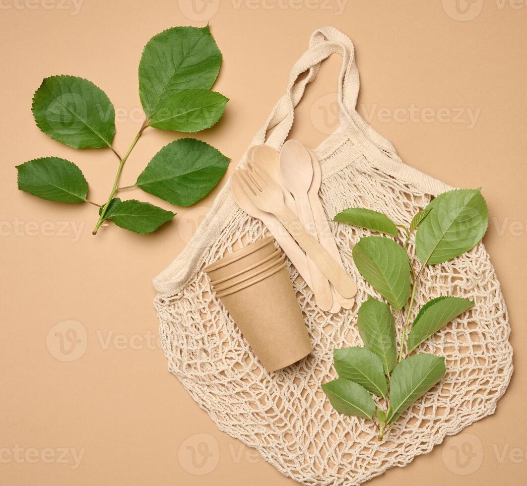 Textile knitted string bag and a stack of disposable cardboard glasses made of brown paper on a beige background and green leaves, top view photo