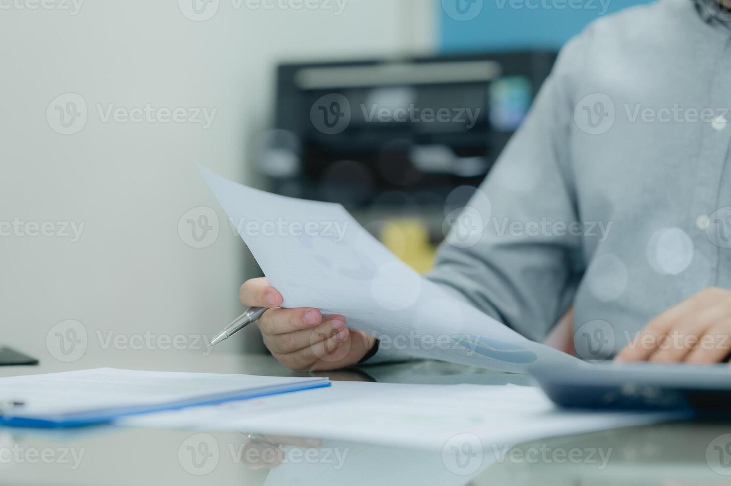A man is sitting at a desk with a piece of paper in his hand. He is looking at the paper and he is focused on it. The desk is cluttered with various items, including a calculator, a pen, and a laptop photo