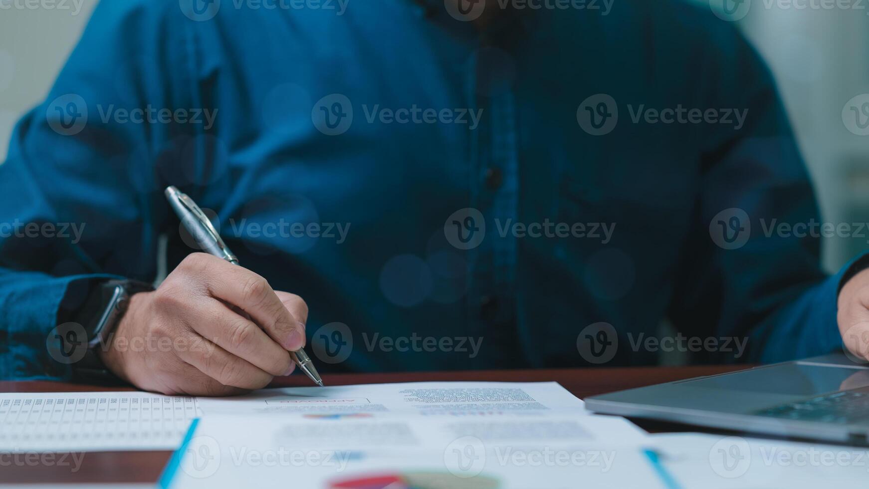 Businessperson is writing on a piece of paper with a pen. Concept of focus and concentration as the man carefully writes. The blue shirt he is wearing adds a touch of professionalism to the scene photo