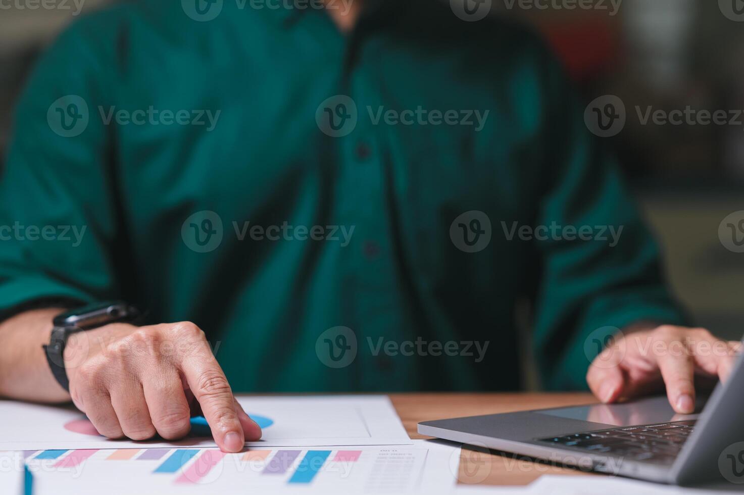 Marketing manager is sitting at a desk with a laptop and a stack of papers. He is pointing at a graph on the papers. Scene is focused and serious, as the man is analyzing the data on the graph photo