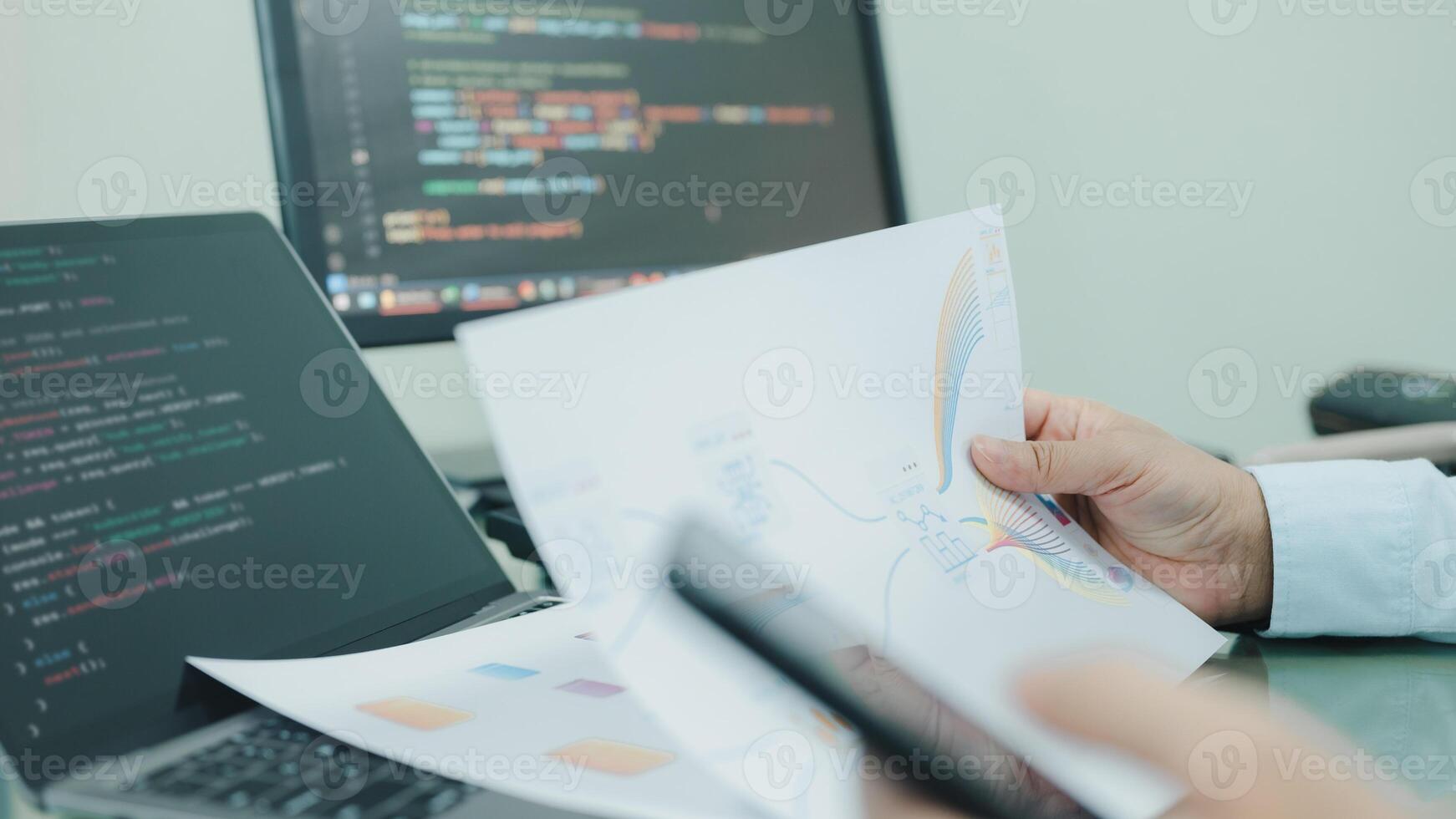 A programmer is holding a piece of paper and a cell phone, possibly working on a project or reading something. The scene suggests a focus on technology and productivity photo