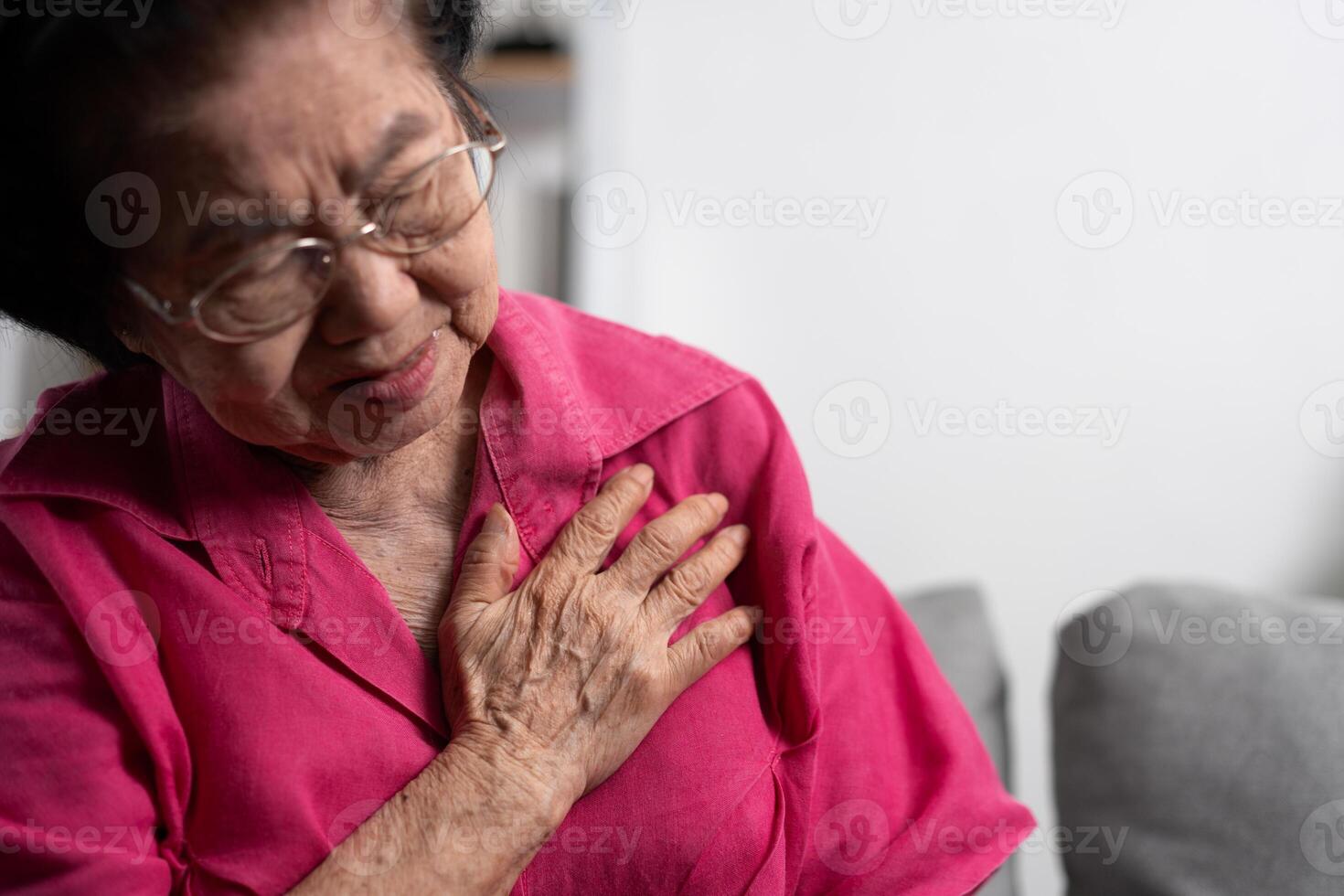 asiático mayor mayor abuela sufrimiento desde corazón dolor y participación su cofre mientras sentado en el sofá en su vivo habitación. médico emergencia tratamiento y salud cuidado seguro proteccion concepto foto