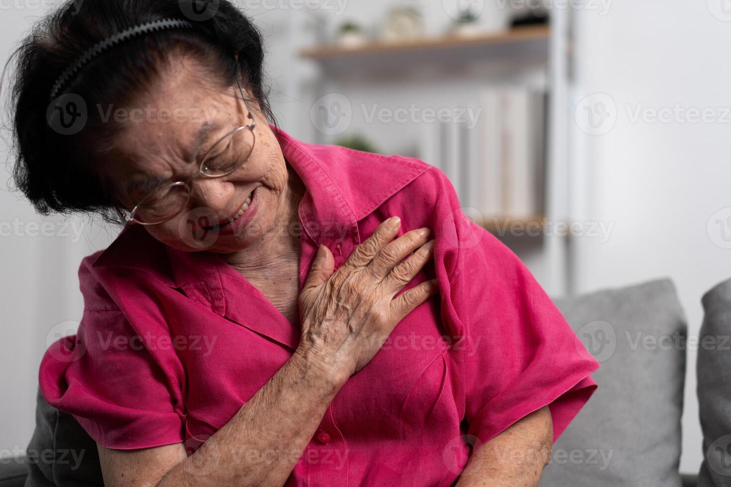 asiático mayor mayor abuela sufrimiento desde corazón dolor y participación su cofre mientras sentado en el sofá en su vivo habitación. médico emergencia tratamiento y salud cuidado seguro proteccion concepto foto