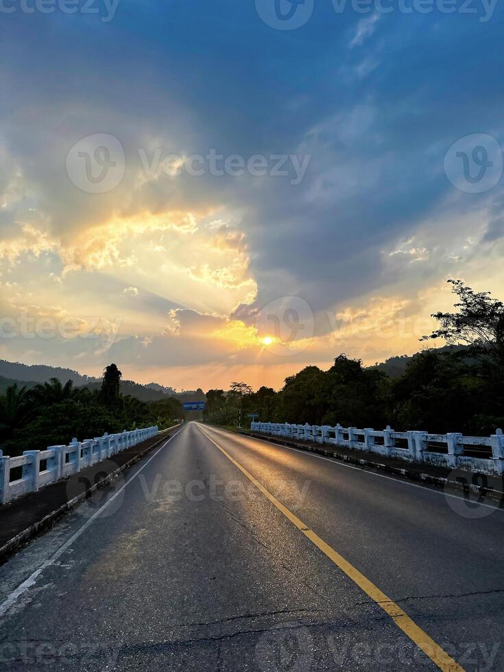 A tranquil highway leads towards a stunning sunset, flanked by lush greenery and radiant skies at dusk. photo