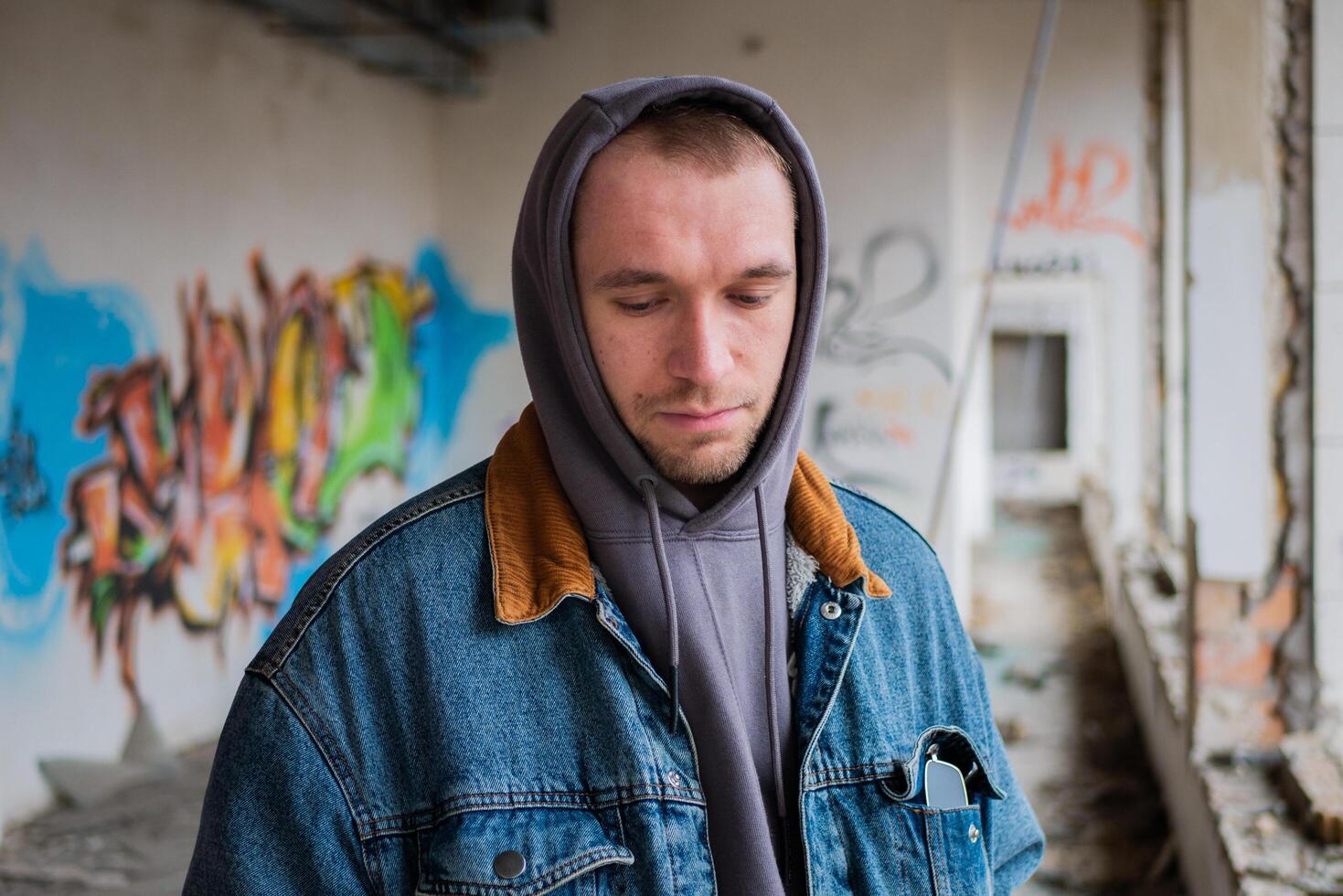 retrato de joven hermoso chico en un mezclilla chaqueta y un gris capucha poses en contra el antecedentes de un destruido y abandonado edificio con pintada. urbano concepto. calle estilo de vida. foto