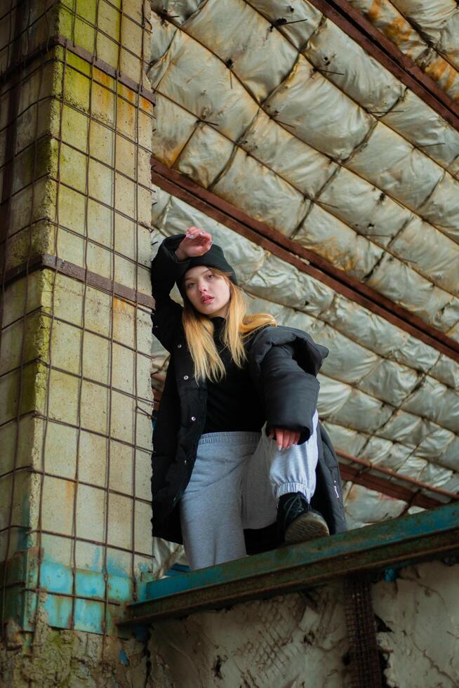 Beautiful teenage girl in a black winter jacket and hat poses in destroyed and abandoned building. Urban concept. Vertical shot. photo