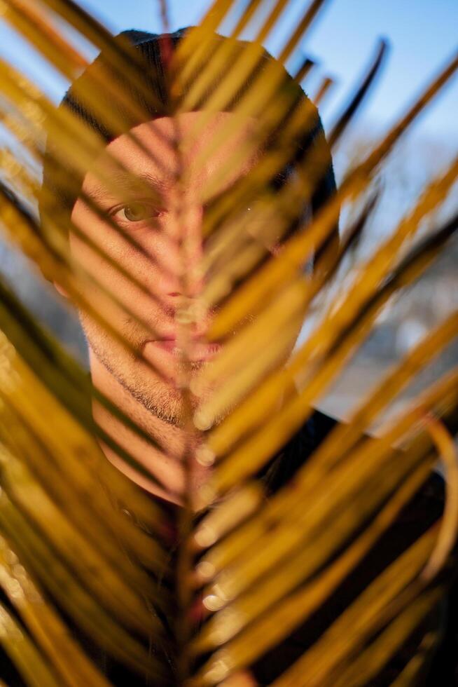 Portrait of a young handsome guy in a black hat covering his face with dry palm leaves. photo