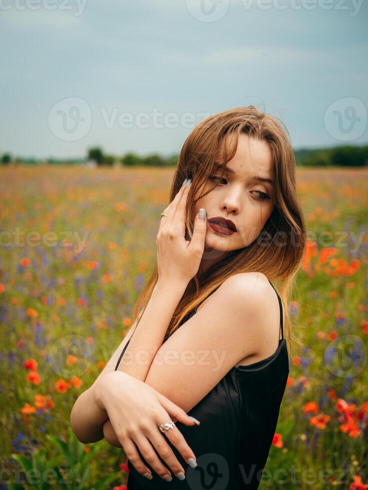 Beautiful young girl in a black evening dress posing against a poppy field on a cloudy summer day. Portrait of a female model outdoors. Rainy weather. Gray clouds. photo