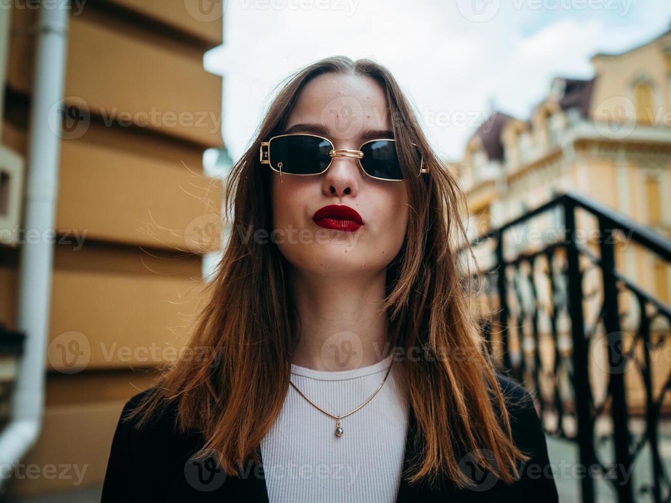 A woman in sunglasses, a white shirt and a black jacket stands confidently on the street. photo