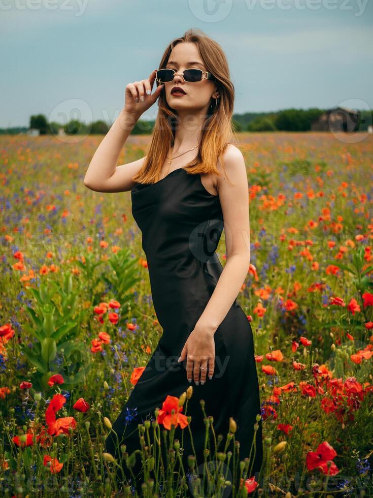 Beautiful young girl in a black evening dress and sunglasses posing against a poppy field on a cloudy summer day. Portrait of a female model outdoors. Rainy weather. Gray clouds. photo