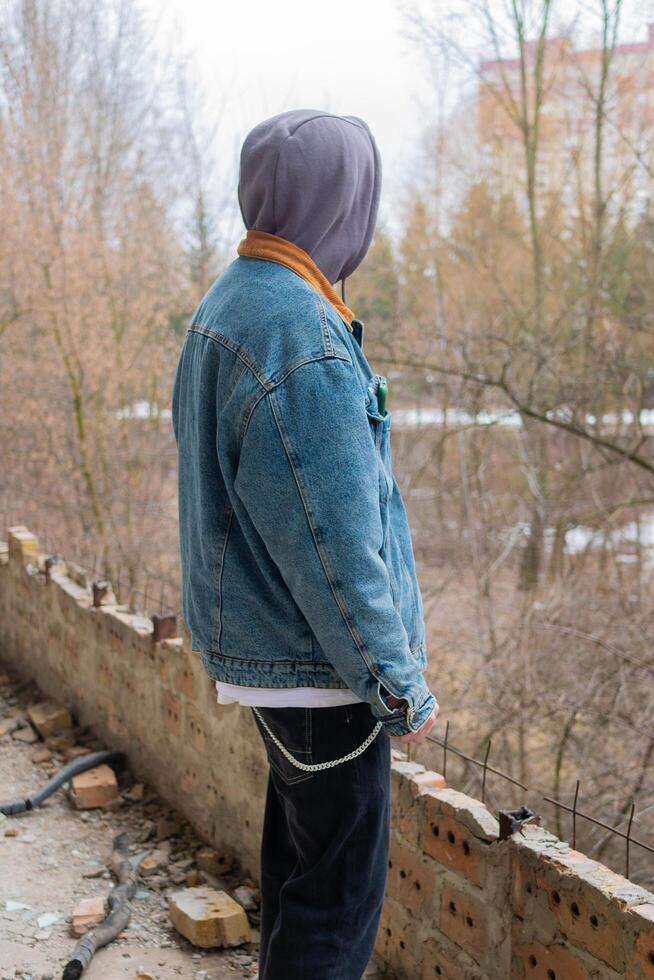 A handsome guy in a denim jacket and a gray hood poses against the background of a destroyed and abandoned building and dry trees without leaves. Cloudy weather. Urban concept. Street lifestyle. photo