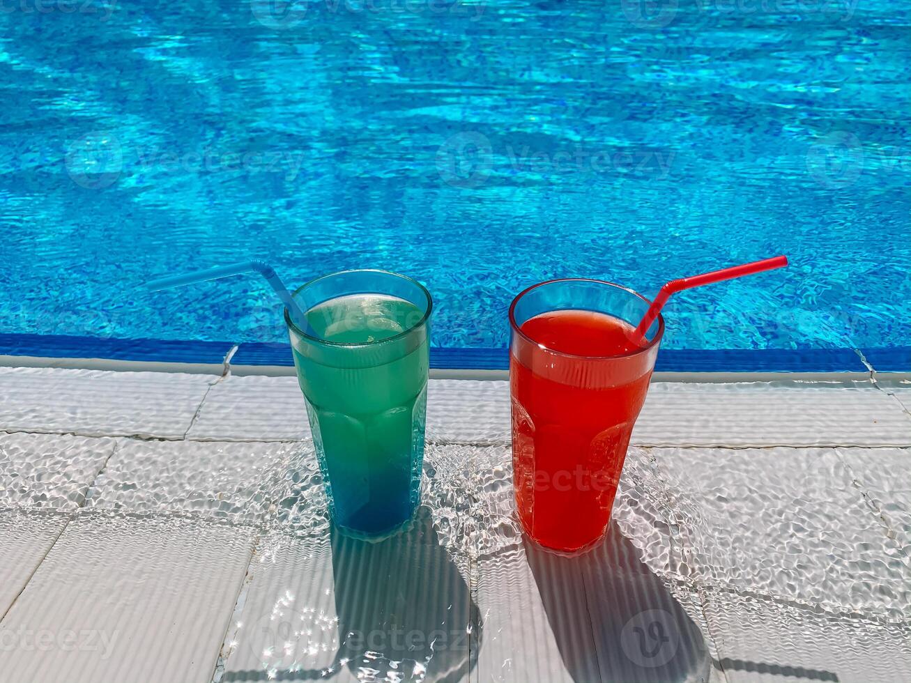 Blue and red cocktails with ice stand on side pool, against background of pool, blue water. Summer pool party, vacation photo