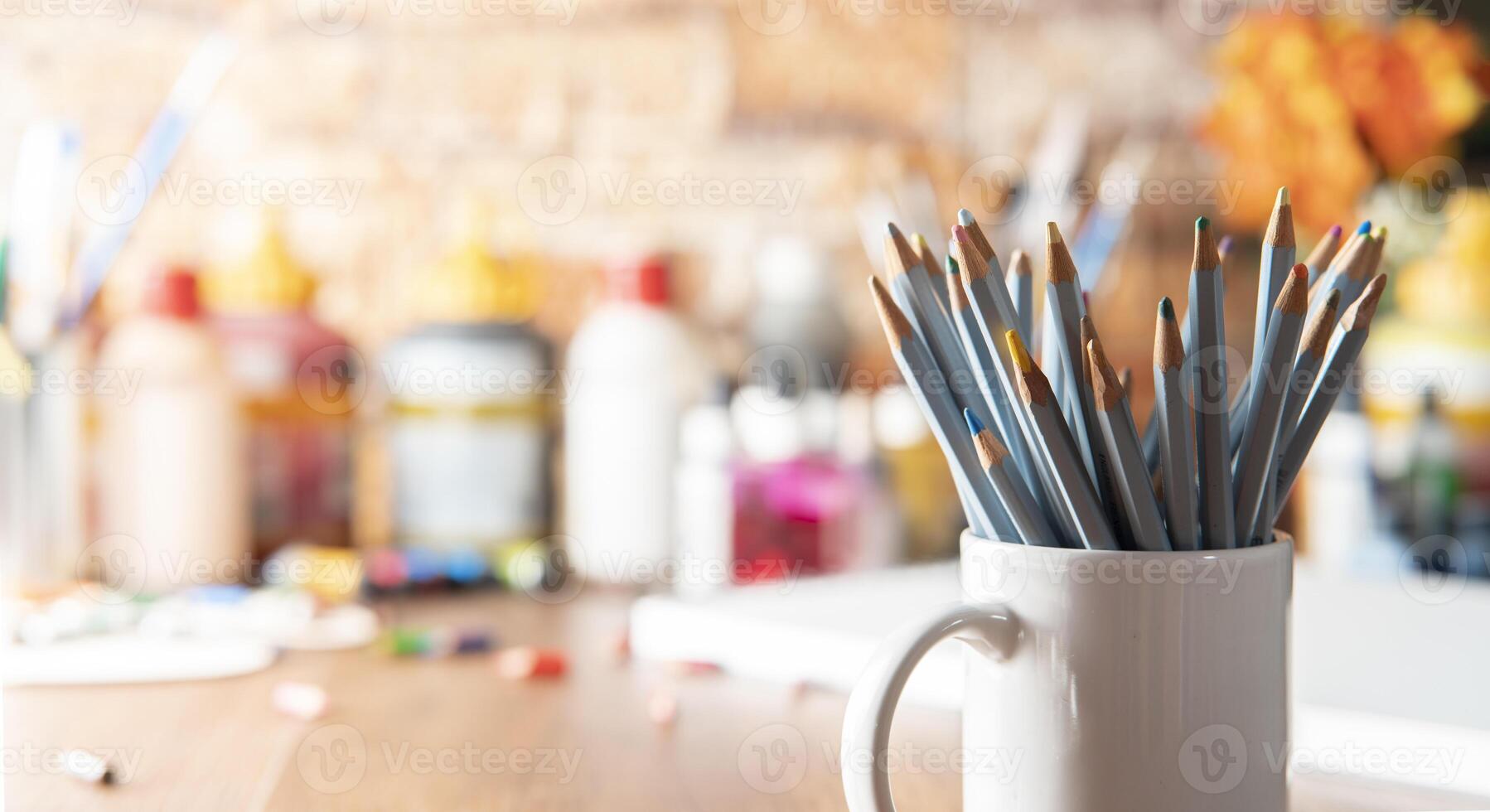 White mug with colored pencils in the foreground on a wooden table with art supplies in the background photo