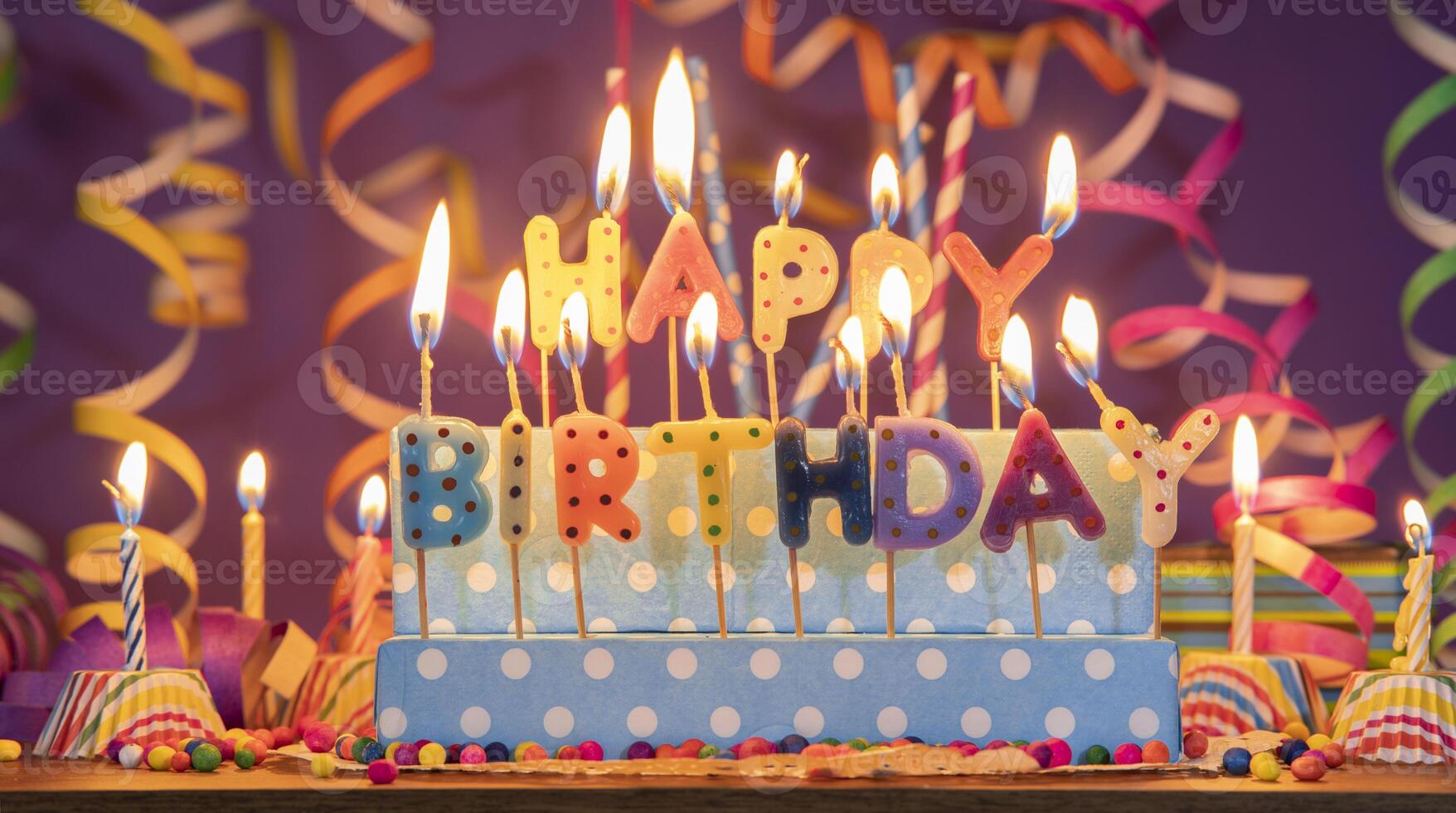 Lit candles of different colors in the shape of letters forming the phrase HAPPY BIRTHDAY, against a purple out-of-focus background photo