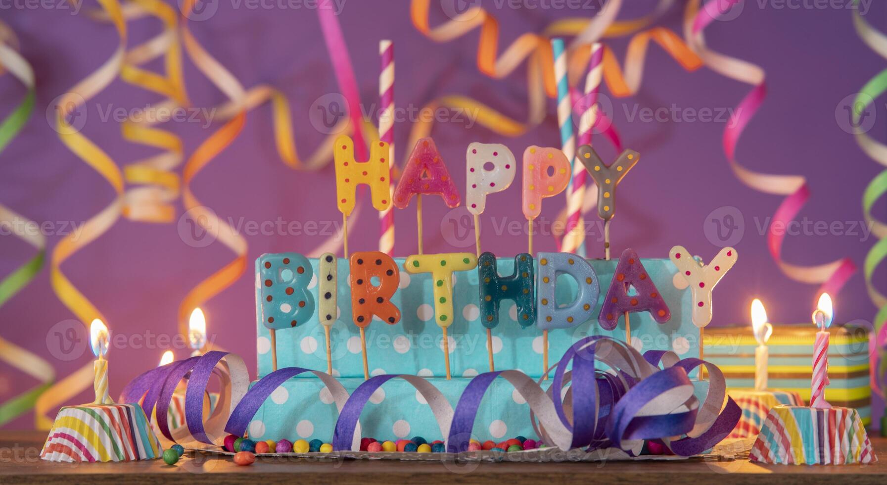 Letters of different colors forming the phrase HAPPY BIRTHDAY, with lit candles against purple out-of-focus background photo