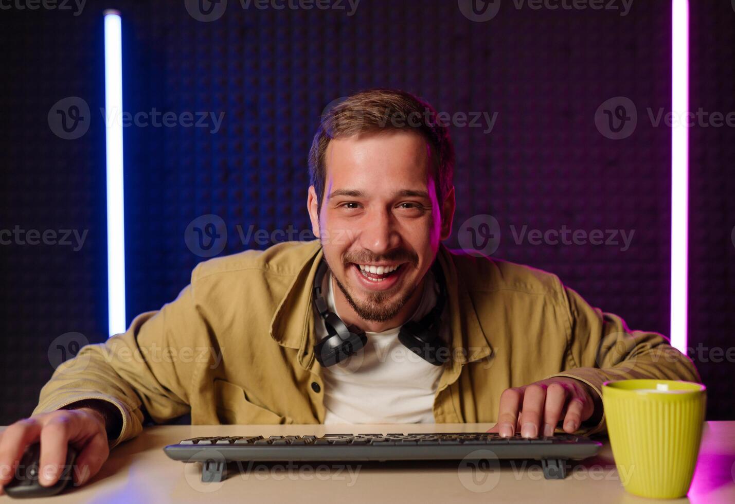 Handsome man in shirt clothes and headset sitting at a table in a room with neon lights and playing games on the computer with a smile on his face looking at the screen. photo