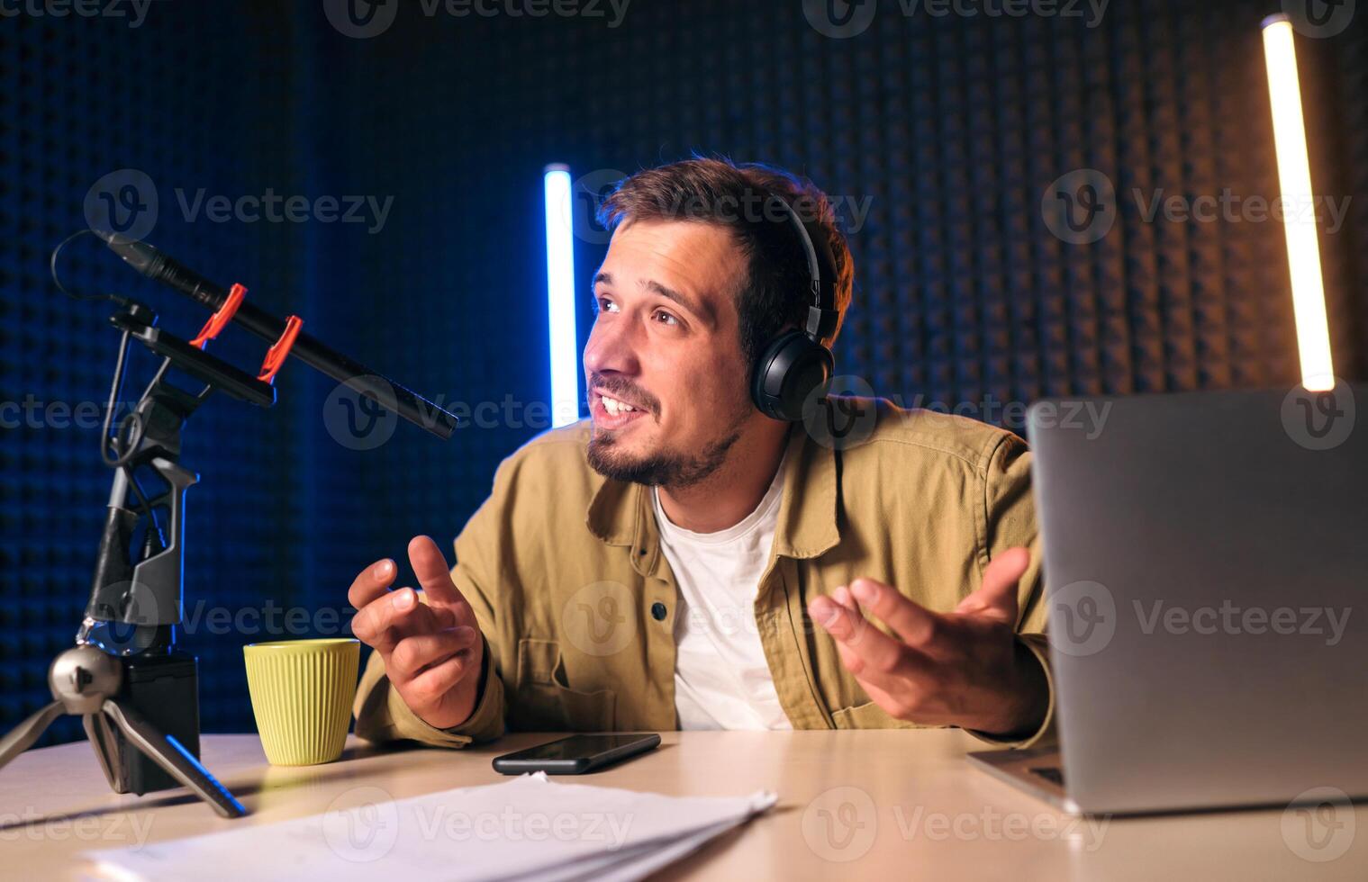 Young stylish man in mustard shirt with headphones gesturing at microphone and sharing story with audience while sitting at desk in studio with neon lighting and recording podcast photo