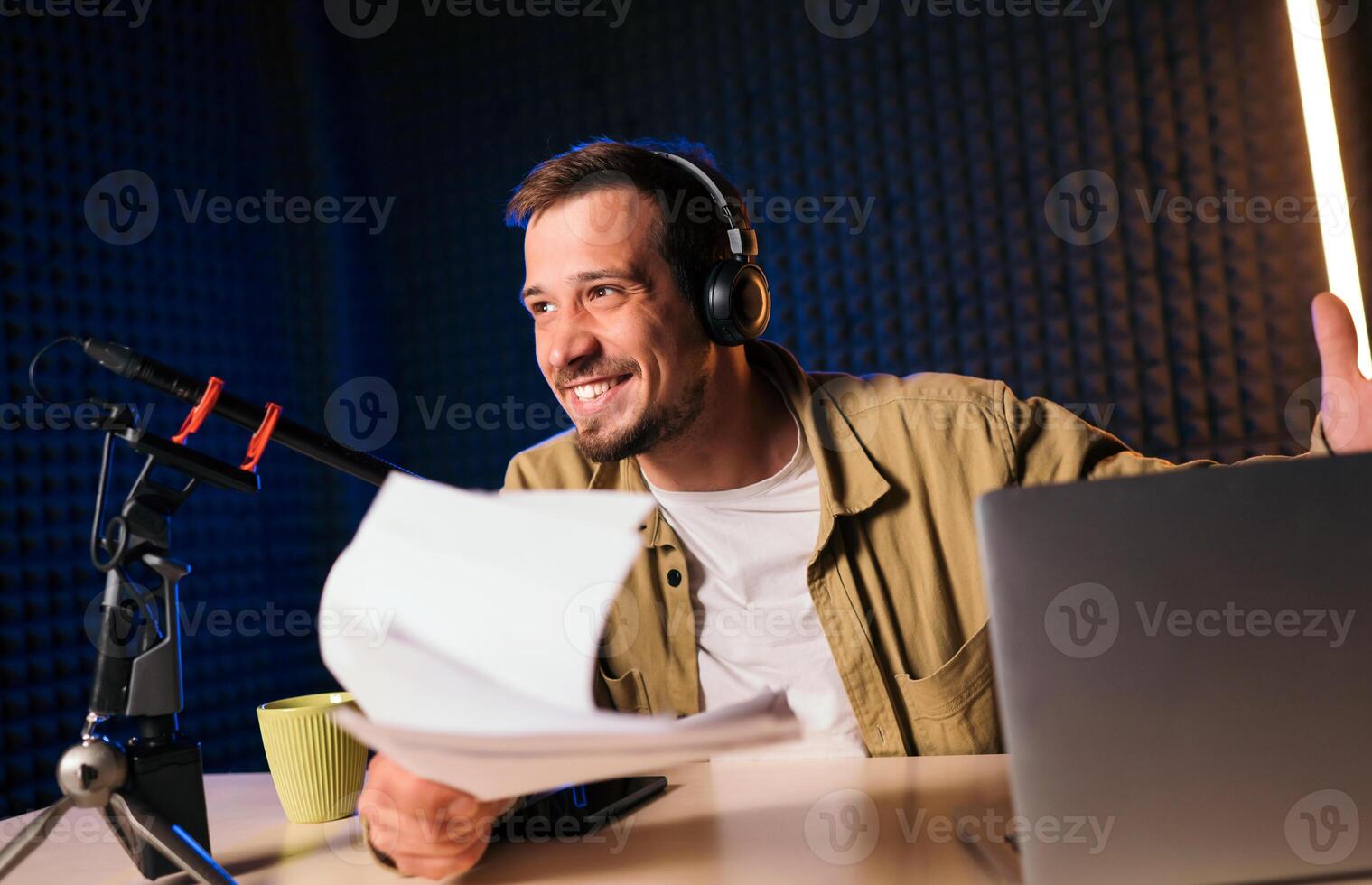 Smiling and gesturing radio host with headphones reading news from paper into studio microphone at radio station with neon lights photo