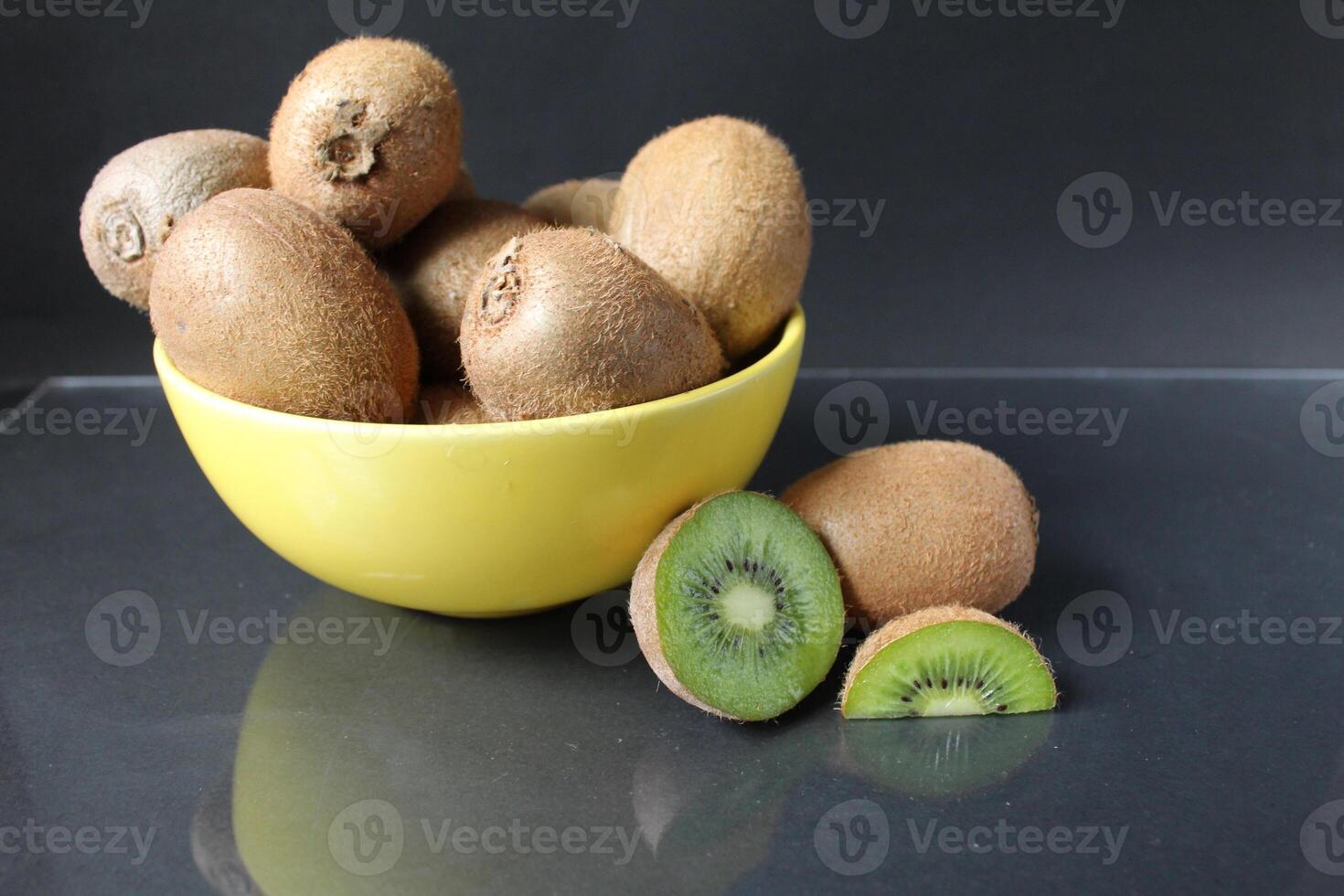 todavía vida de kiwi Fruta en un amarillo cuenco plato y piezas de jarra siguiente a en un negro antecedentes con un reflexión de allí es un copyspace foto