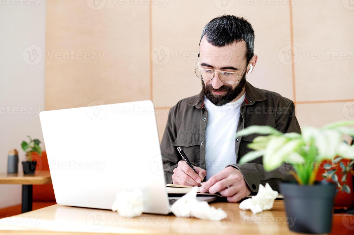 Concentrated man in glasses taking notes writing information from laptop in cafe, cheerful bearded man studying in cafe photo