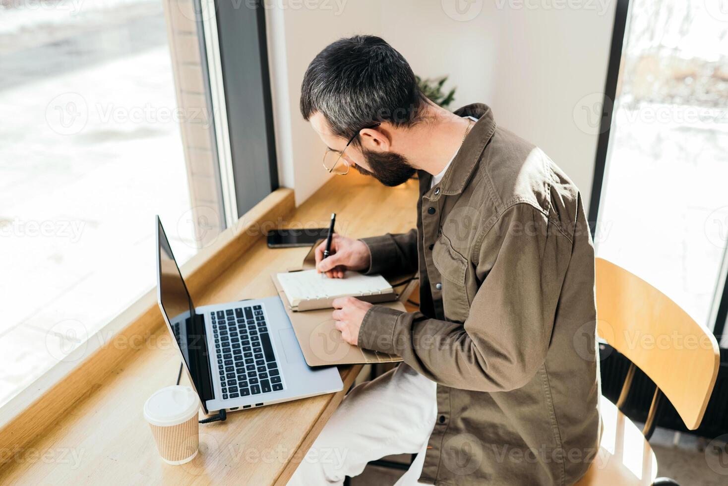 A caucasian man write in notepad on a cafe. work process. A man with a beard in a shirt is sitting in a cafe with a laptop, remote work, freelance photo