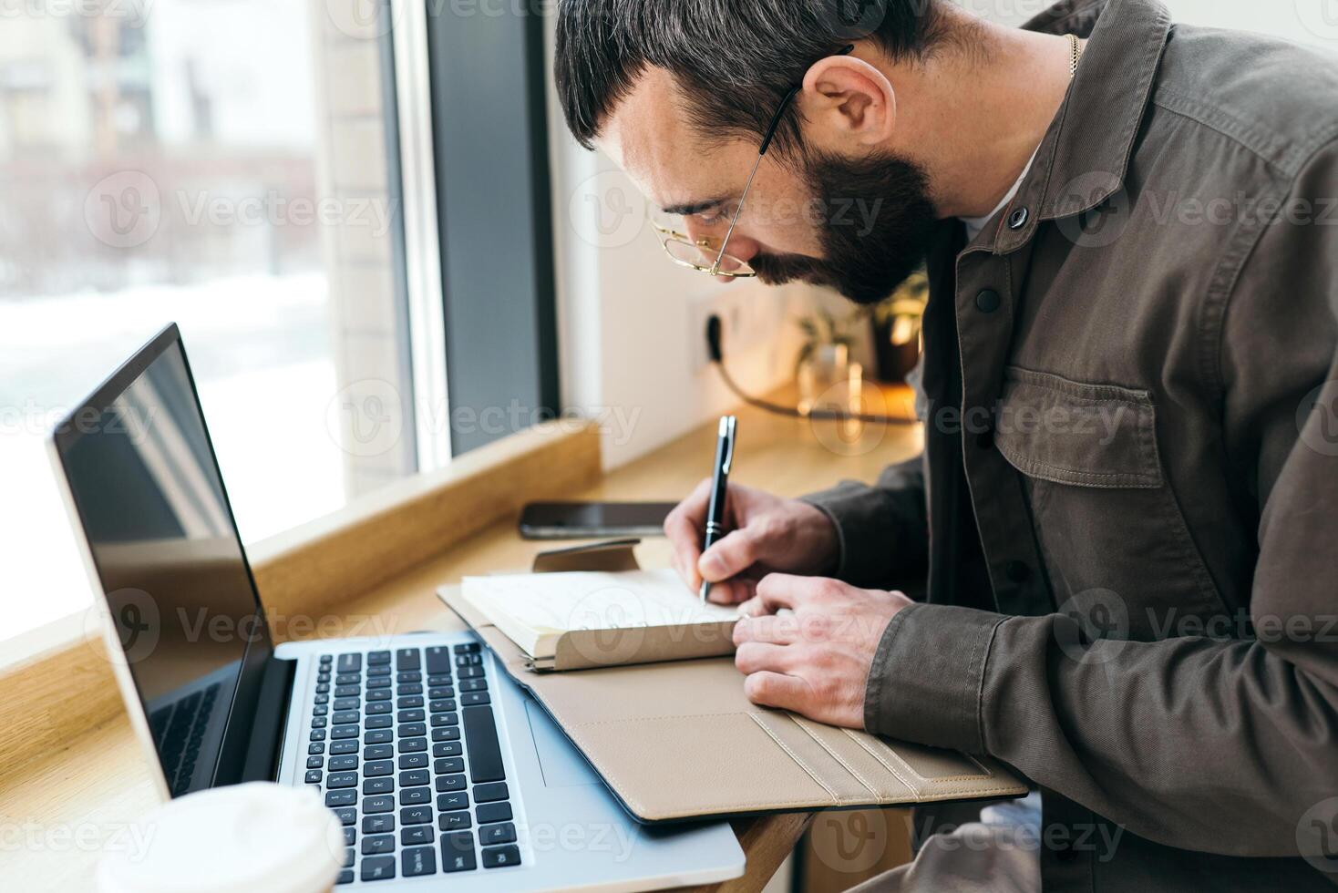 A caucasian man write in notepad on a cafe. work process. A man with a beard in a shirt is sitting in a cafe with a laptop, remote work, freelance photo