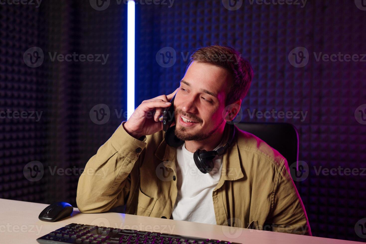 Young stylish man in mustard shirt with headphones gesturing at microphone and sharing story with audience while sitting at desk in studio with neon lighting and recording podcast photo