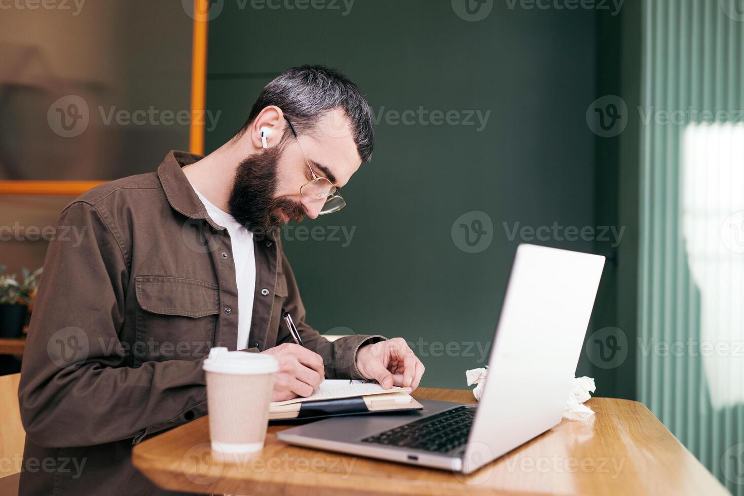 Stylish bearded man sitting at table with laptop and cup of coffee while spending time in modern cafeteria and working on distance photo