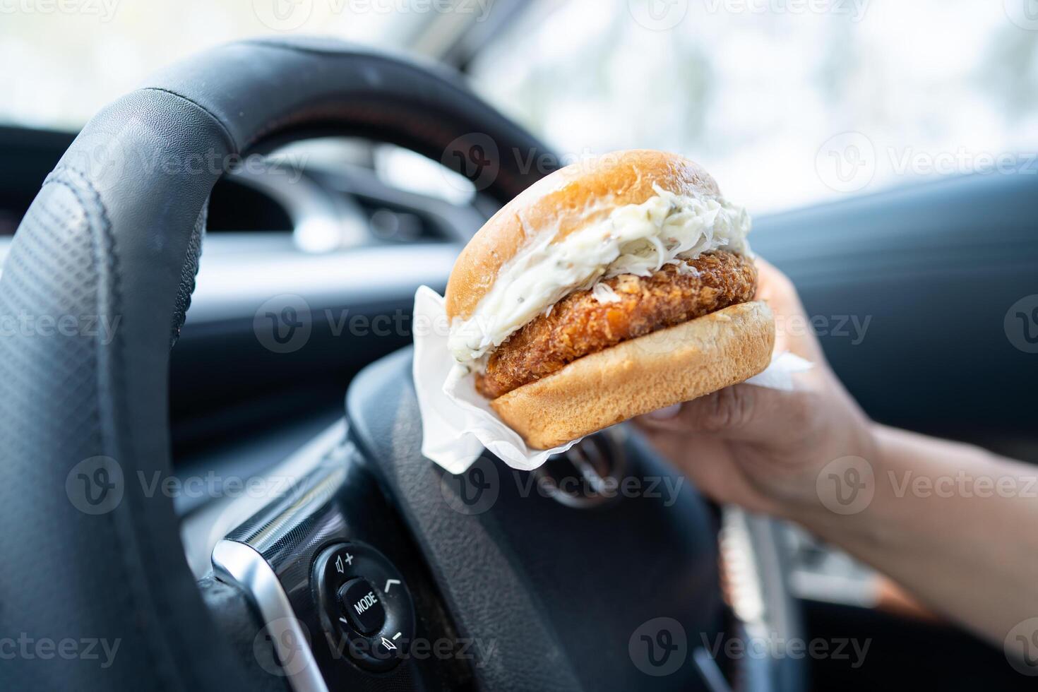 Asian lady holding hamburger to eat in car, dangerous and risk an accident. photo