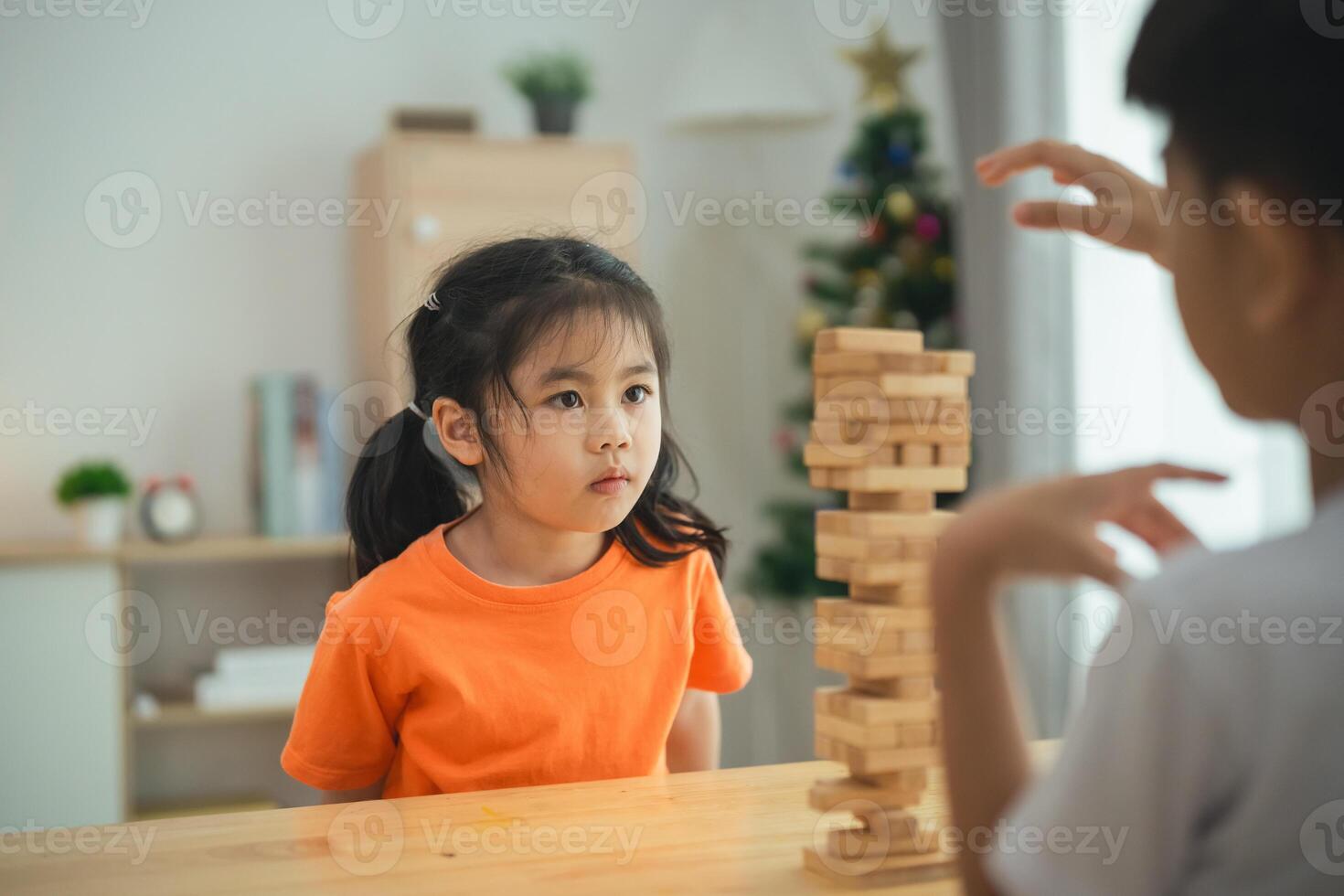 un niña en un naranja camisa es sentado a un mesa con un apilar de de madera bloques un chico es en pie detrás su, acecho su. escena es juguetón y alegre foto