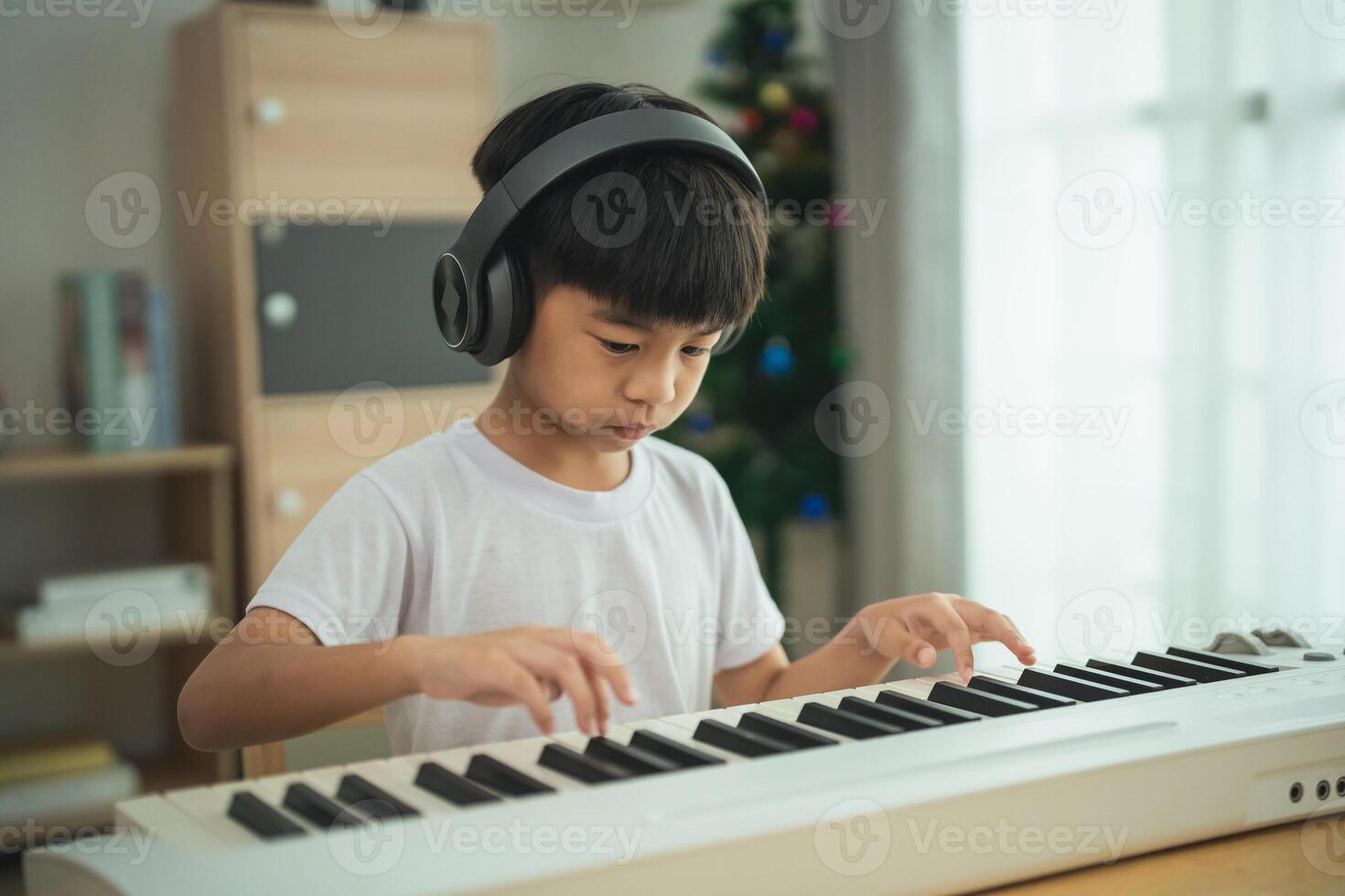 A young boy is playing the piano in a living room. He is wearing headphones and he is focused on the music. The room is decorated with Christmas decorations, including a tree and a few books photo