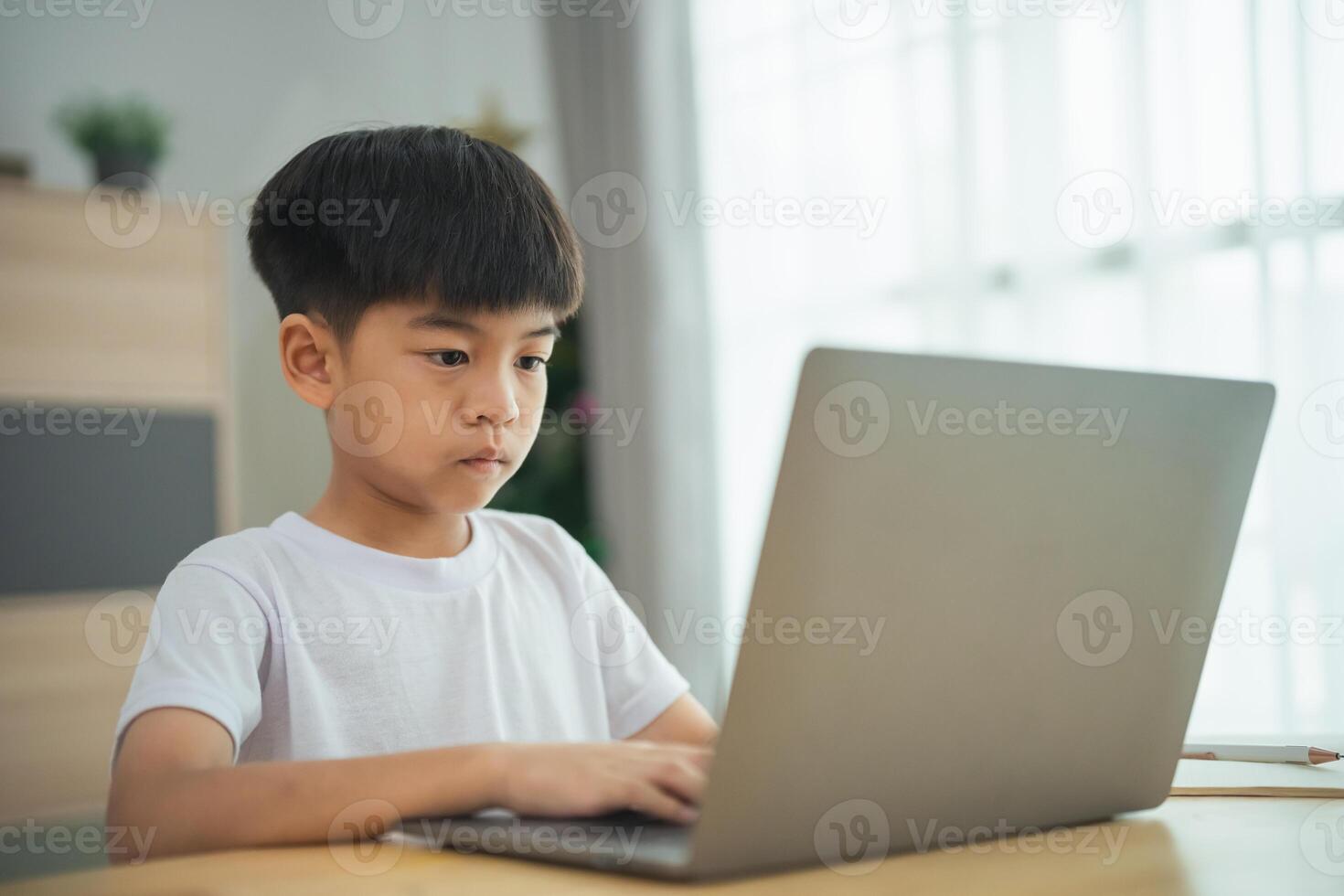 A young boy is sitting at a table with a laptop in front of him. He is focused on the screen, possibly working on a school assignment or playing a game photo