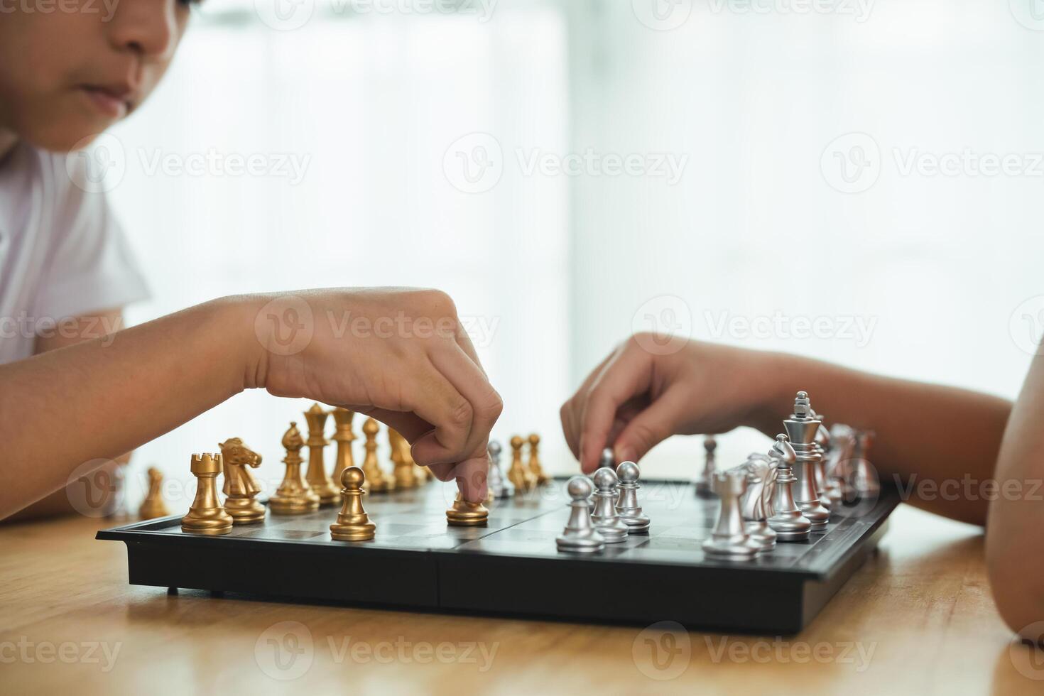 Two children playing a game of chess. One of the children is wearing a white shirt. The game is being played on a black and white board photo