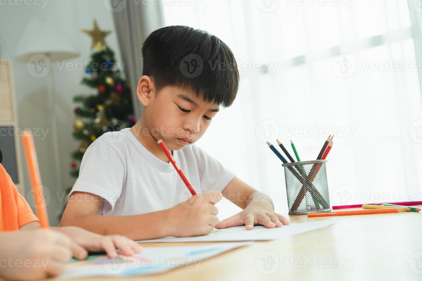 A boy is drawing with a pencil on a piece of paper. He is focused on his drawing and seems to be enjoying himself. The scene is set in a room with a Christmas tree in the background photo