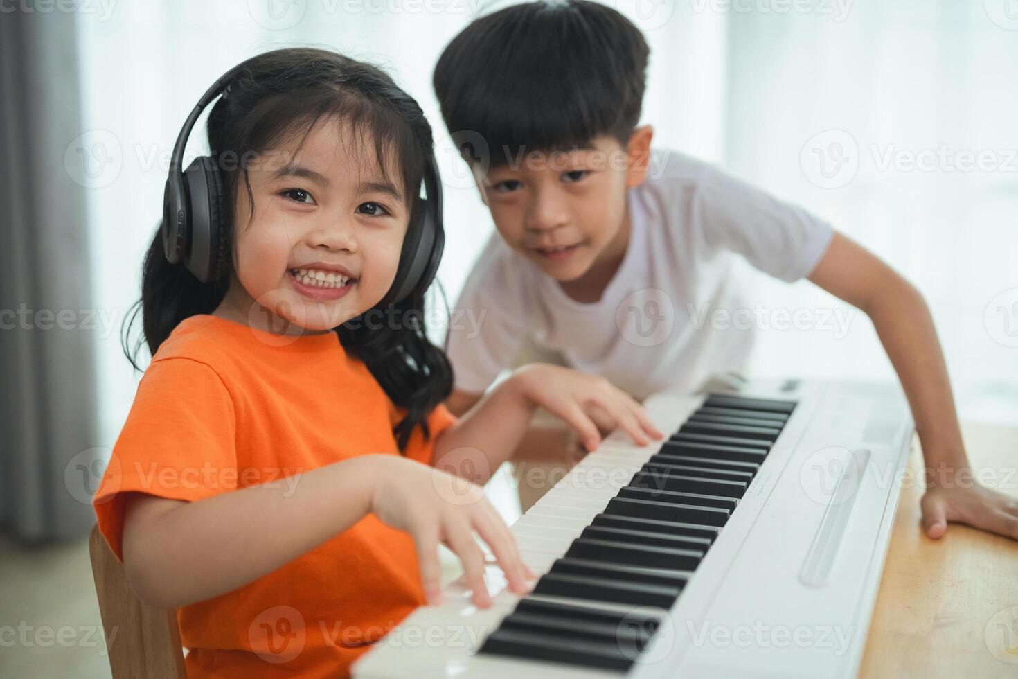 Two children are playing the piano. One is wearing an orange shirt. They are smiling and seem to be enjoying themselves photo