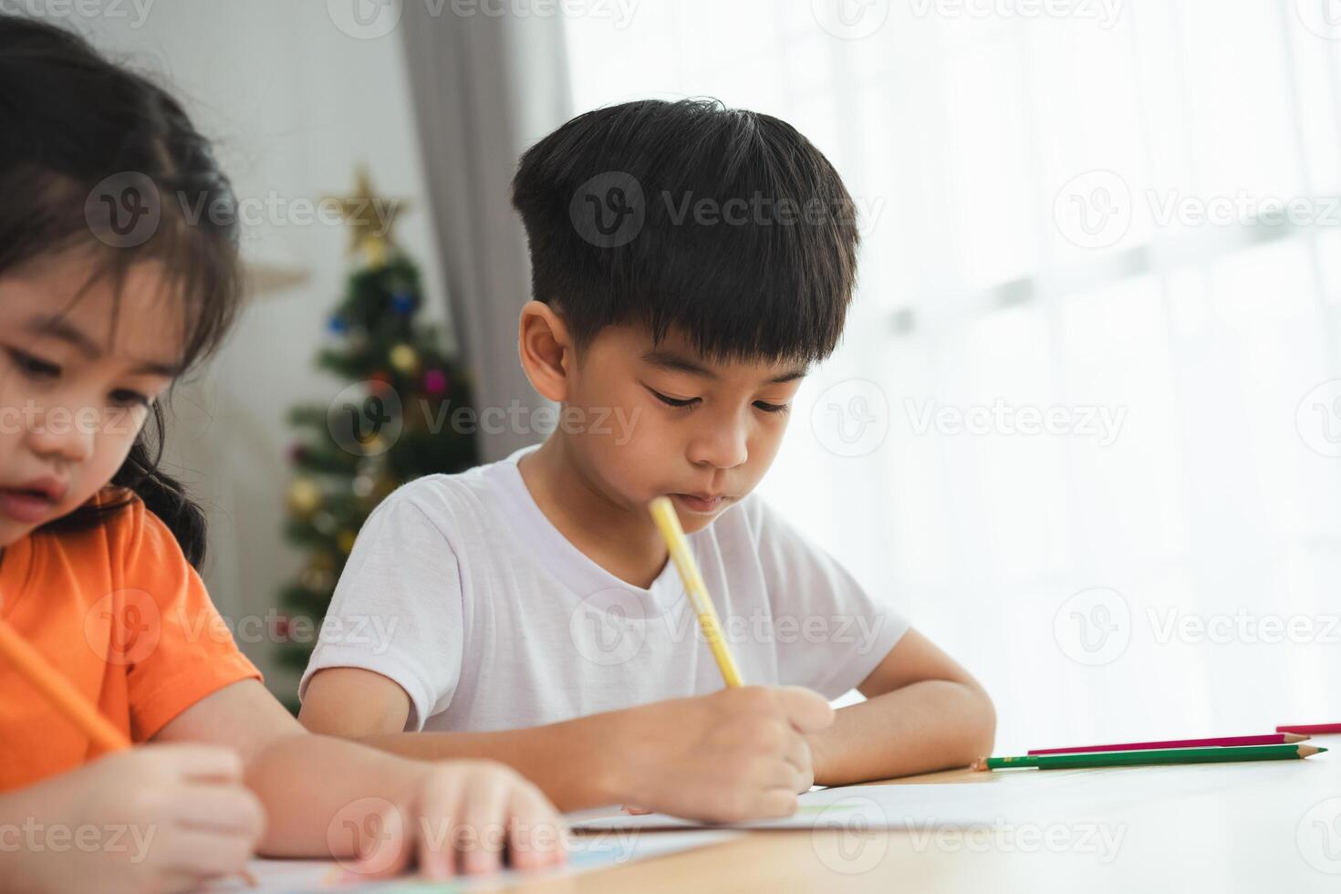un chico es escritura con un lápiz mientras un niña relojes. el niña es vistiendo un naranja camisa foto