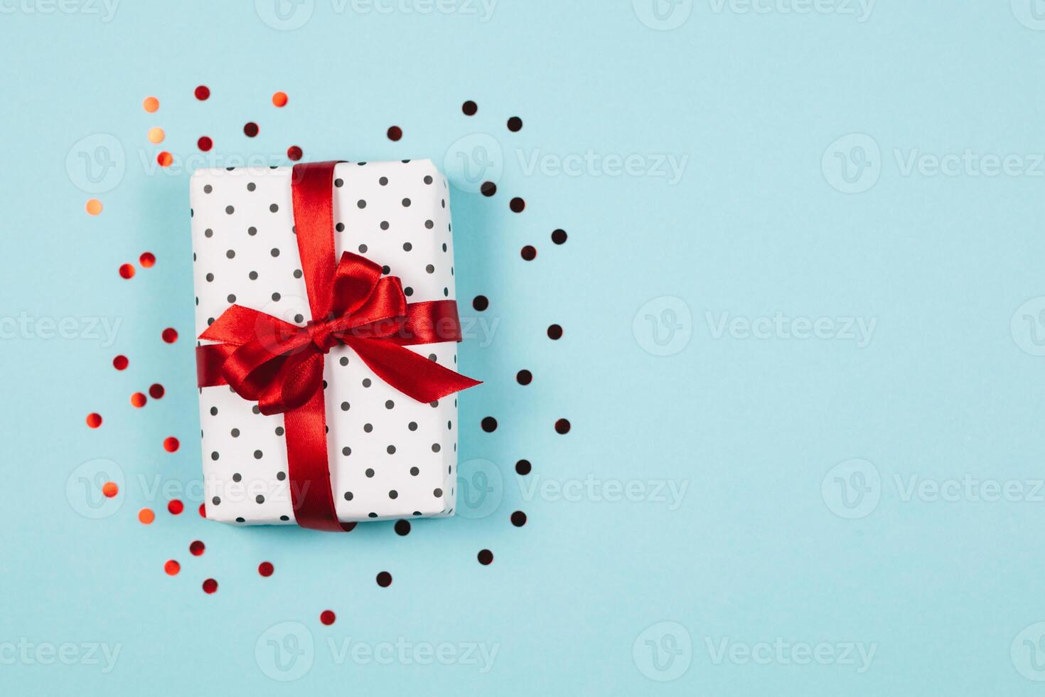 Gift box wrapped in black polka-dot white paper with red bow on blue background with confetti. photo