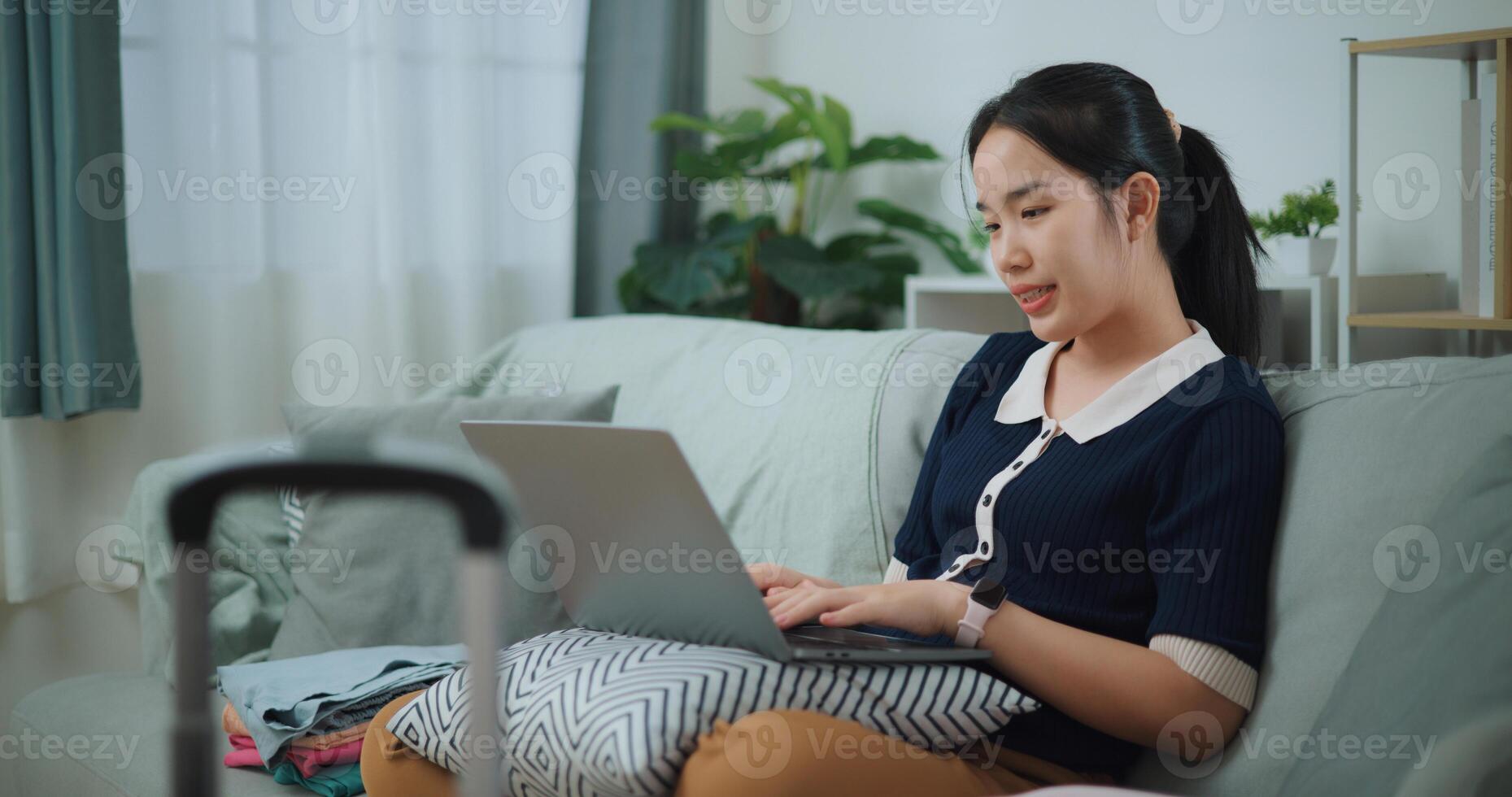 Side view of Asian teenager woman sitting on sofa using laptop for prepare booking hotel and airplane ticket for travel. backpacker travel concept. photo