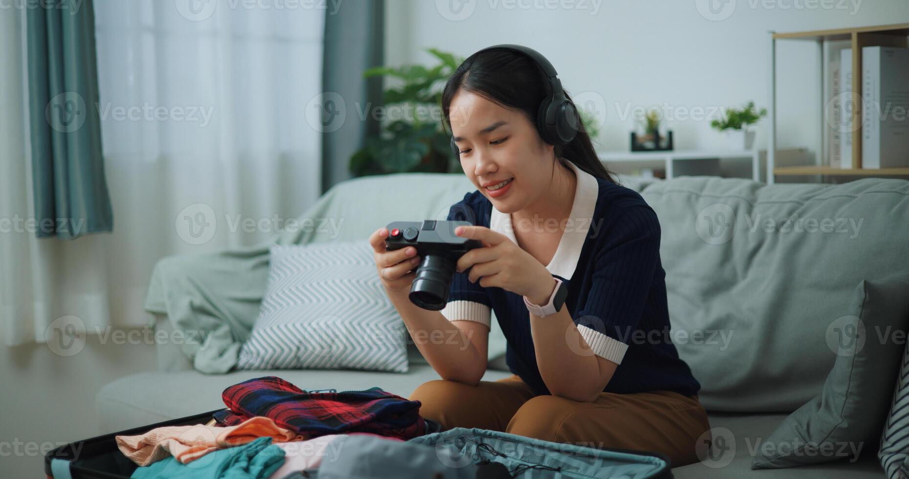 Selective focus of Asian teenager woman wear wireless headphones sitting on sofa look pictures on the camera while packing suitcase luggage in the living room , Preparation travel suitcase at home. photo