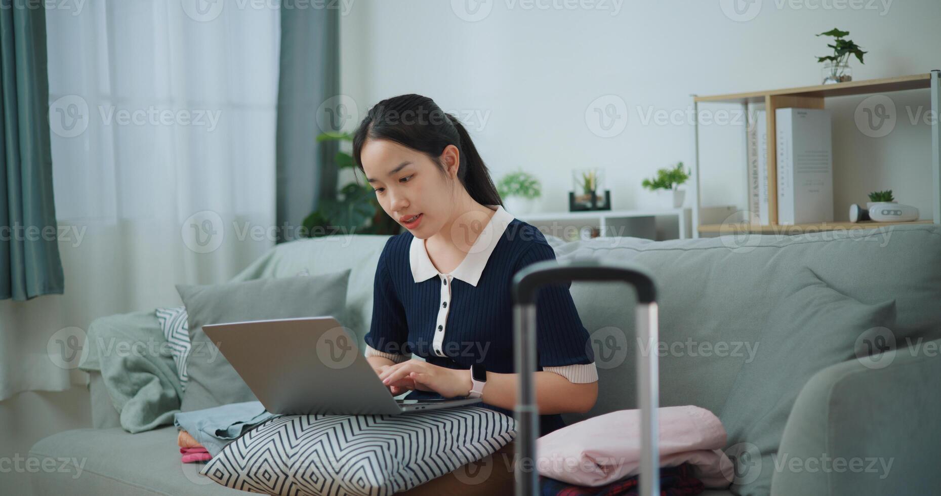 Portrait of Asian teenager woman sitting on sofa using laptop for prepare booking hotel and airplane ticket for travel. backpacker travel concept. photo