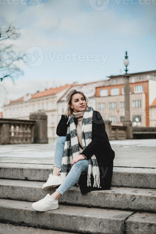 un mujer exuda calor y comodidad como ella se sienta en escaleras, vistiendo un negro Saco y a rayas bufanda. su sonrisa irradia un sentido de contentamiento, atractivo espectadores a compartir en su alegría. foto