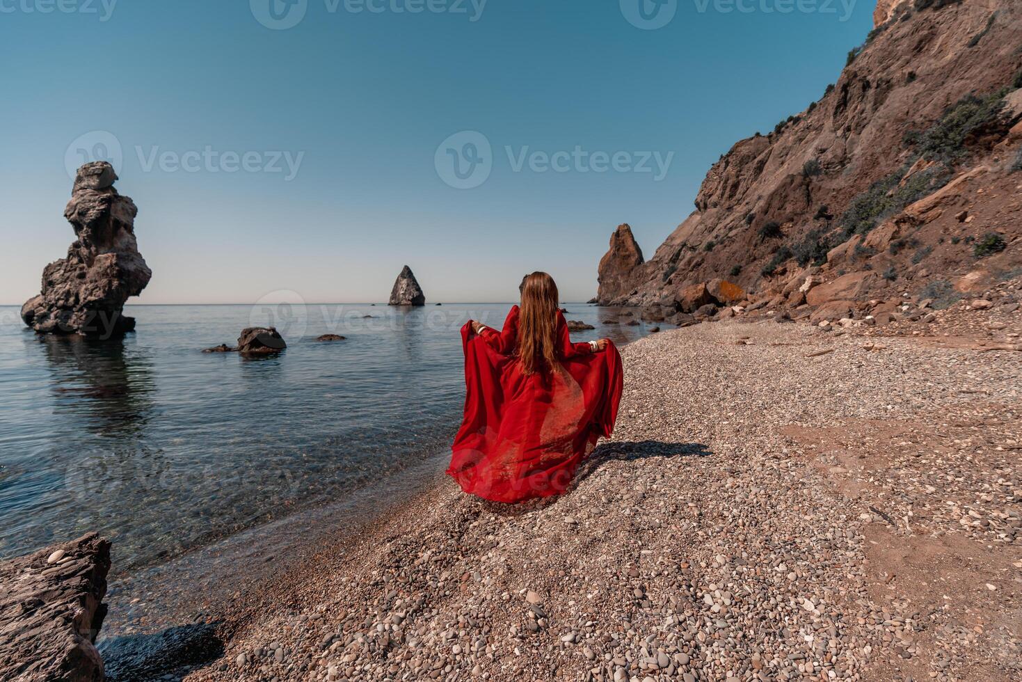 A woman in a red dress stands on a beach with a rocky shoreline in the background. The scene is serene and peaceful, with the woman's red dress contrasting against the natural elements of the beach. photo