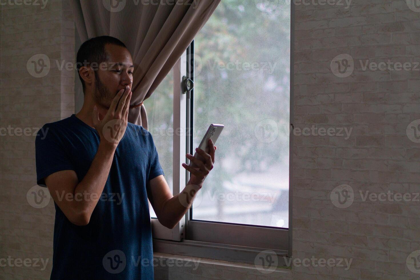 joven asiático hombre en azul camiseta en pie en ventana cerca su boca. obtener sorprendido y sensación contento por su teléfono inteligente foto