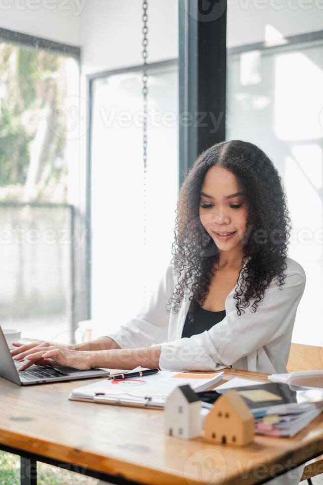 Professional real estate agent is focused on her laptop with house models and documents on the desk, in a bright office. photo